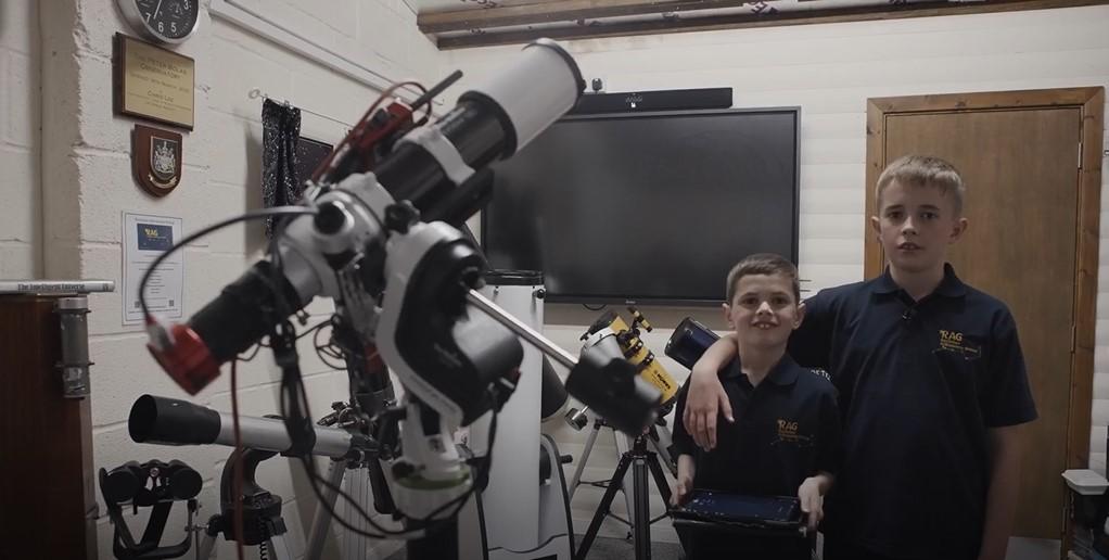 Photo of two young brothers standing next to each other and smiling at the camera, the older brother has his arm around the younger. On the left is a large complex telescope set up with wires