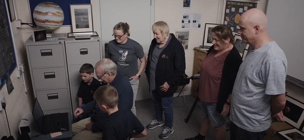 Photo of group of people including two children who are sitting down looking at a computer screen, standing in a room with astronomy decorations like a model of Jupiter