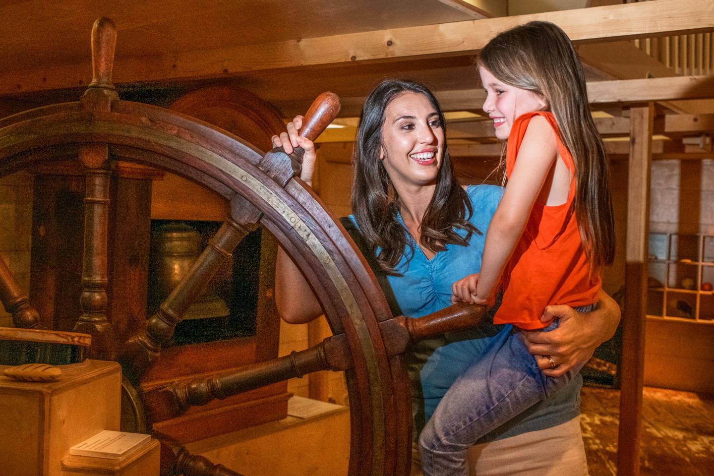 A mother and daughter play in the All Hands Children's Gallery at the National Maritime Museum. Mum has one hand on a large wooden ship's steering wheel, and is lifting her daughter up with her other arm