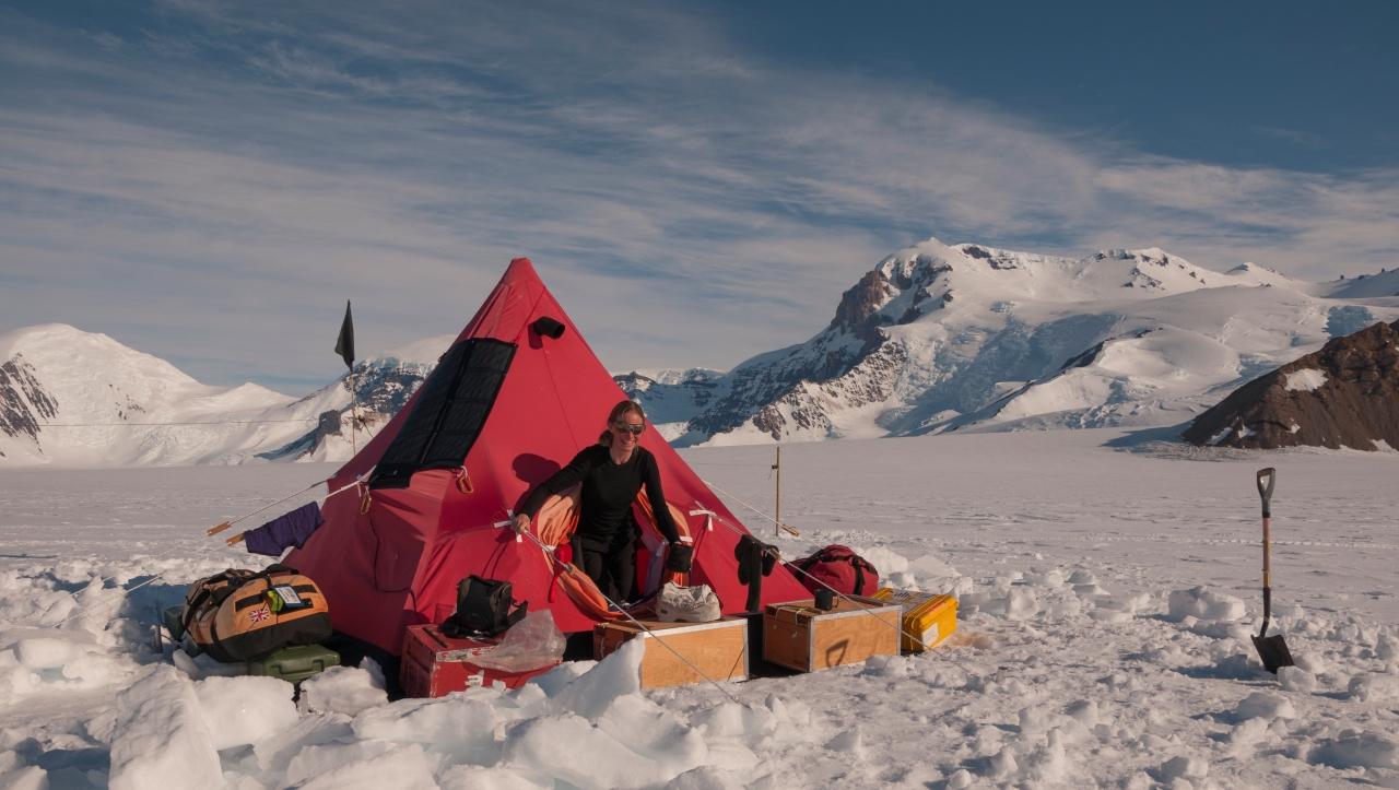 A scientists looks out from a red pyramid tent in Antarctica, with equipment and supplies dotted around the snow