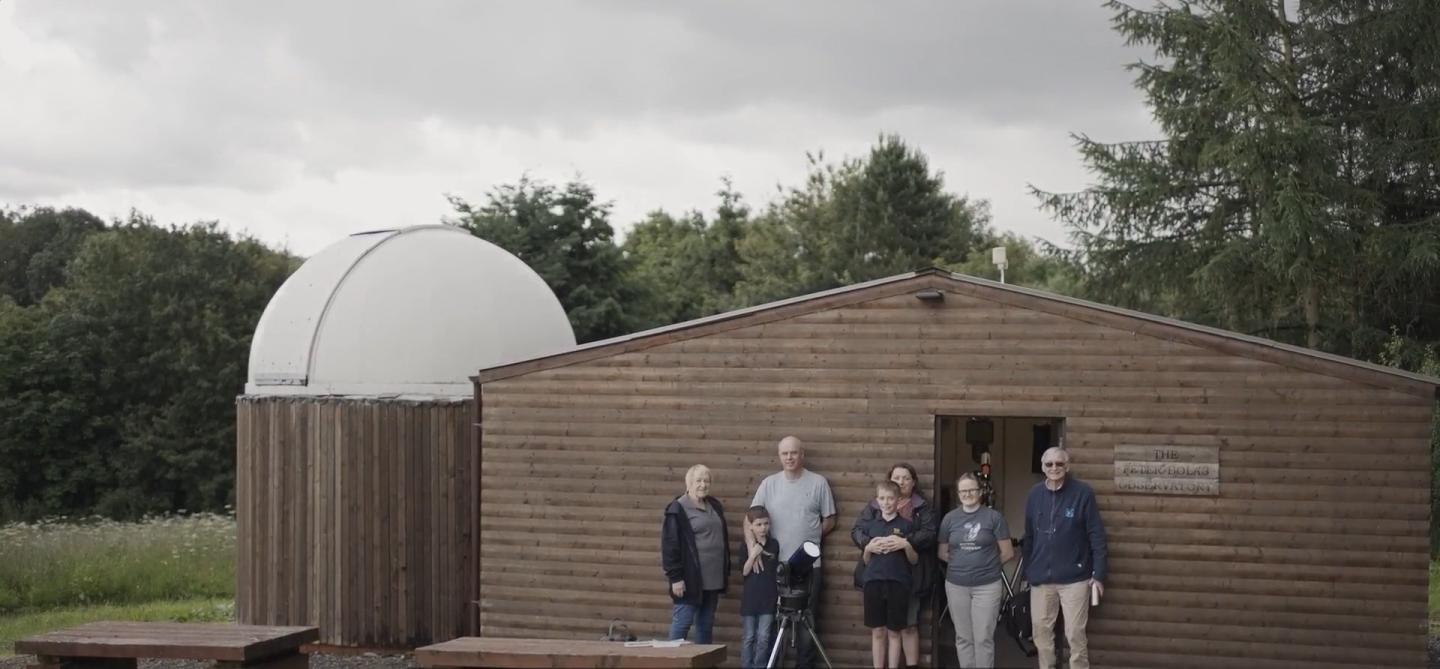 Photo of group of people (some younger and some older) standing outside a wooden hut, next to which is a small onion dome observatory