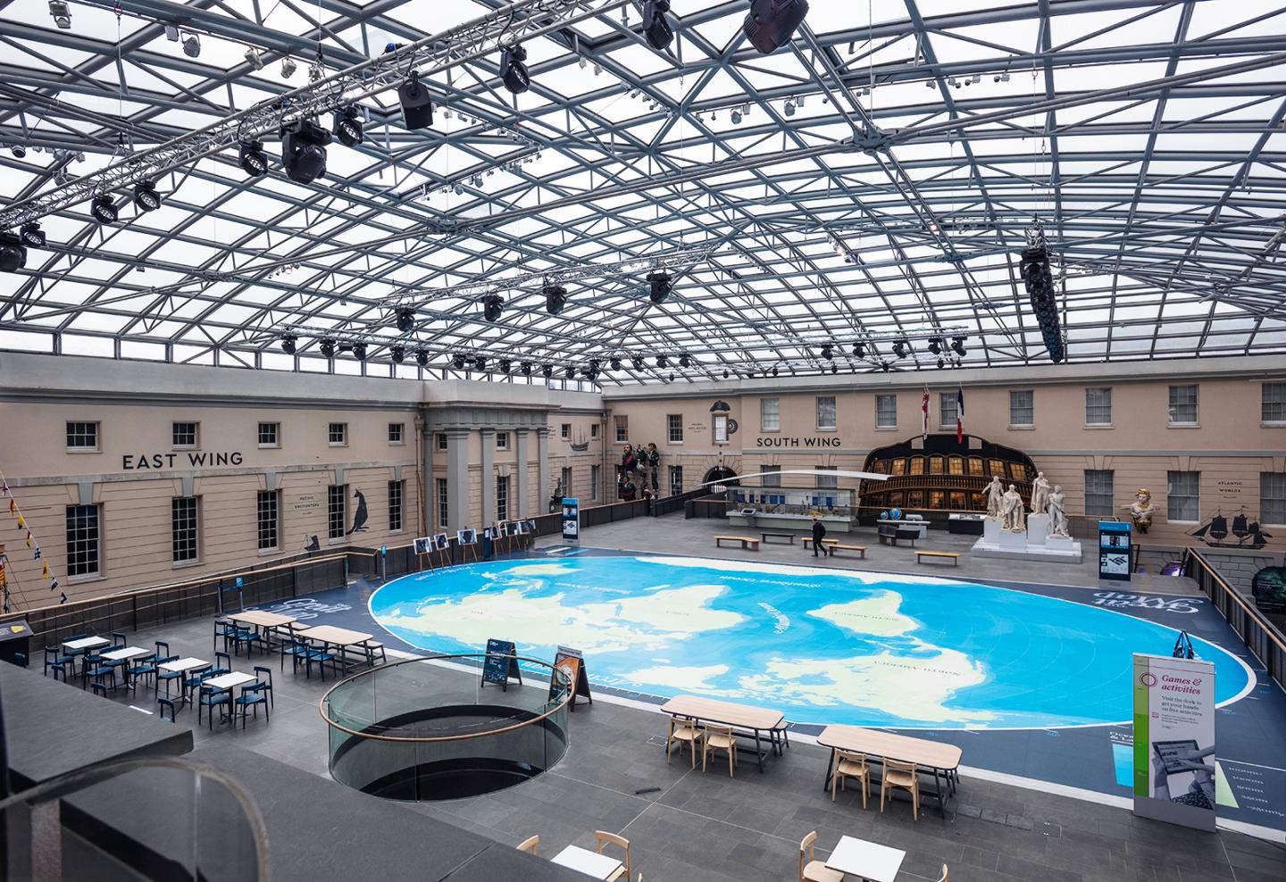 Interior view of the National Maritime Museum, showing a large open space with a giant map on the floor. A glass roof with metal beams stretches across the entire space