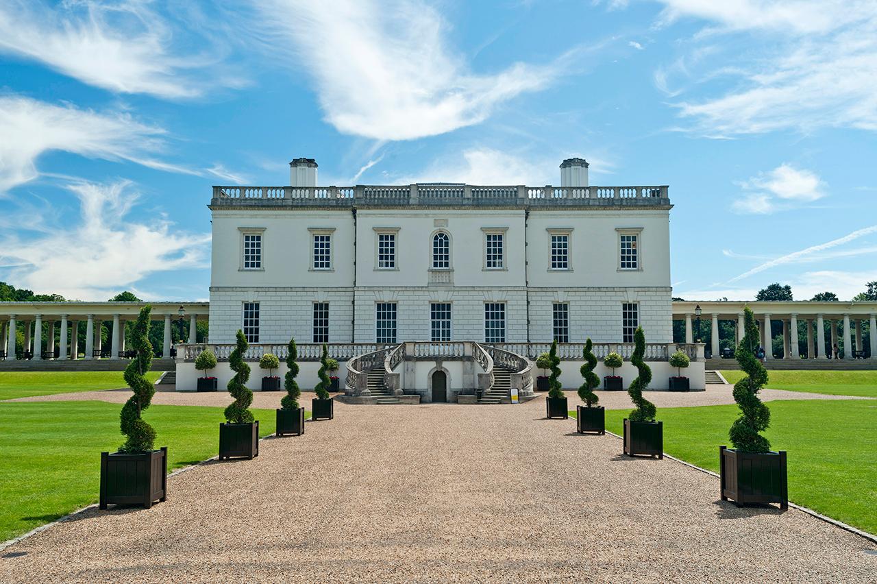 A view of the Queen's House in Greenwich from the outside. The square white building is almost symmetrical from the front, with a gravel pathway leading up to the main entrance